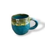 Textured teal ceramic mug with a flowing golden and earthy glaze around the rim and handle, resembling molten metal over rugged terrain.