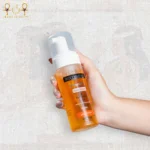 Vitamin C face wash with hyaluronic acid in a 150ml foam pump bottle, bright orange liquid, held in a hand against a textured background.