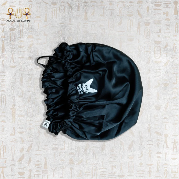 Black satin sleeping bonnet with adjustable strap on neutral background.