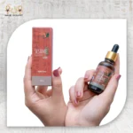 Hands holding Queen sesame essential oil bottle and packaging box against a light fabric background.